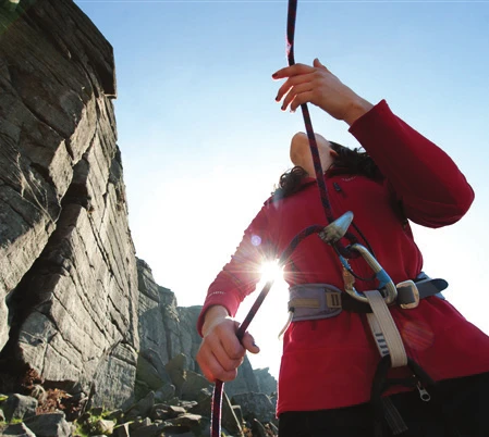 A participant pulling rope through a belay device at the base of a cliff, backlit by low sun