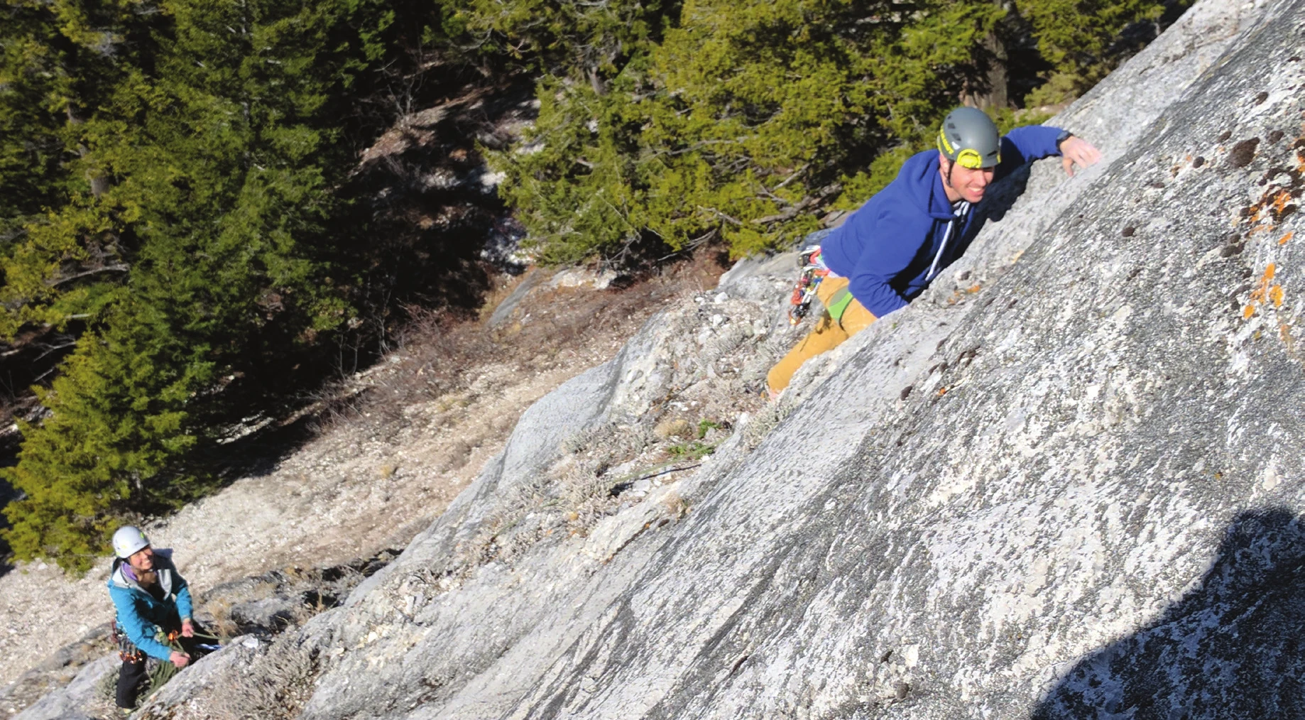 A climber moving up a low-angle slab on a top rope, with the belayer visible below
