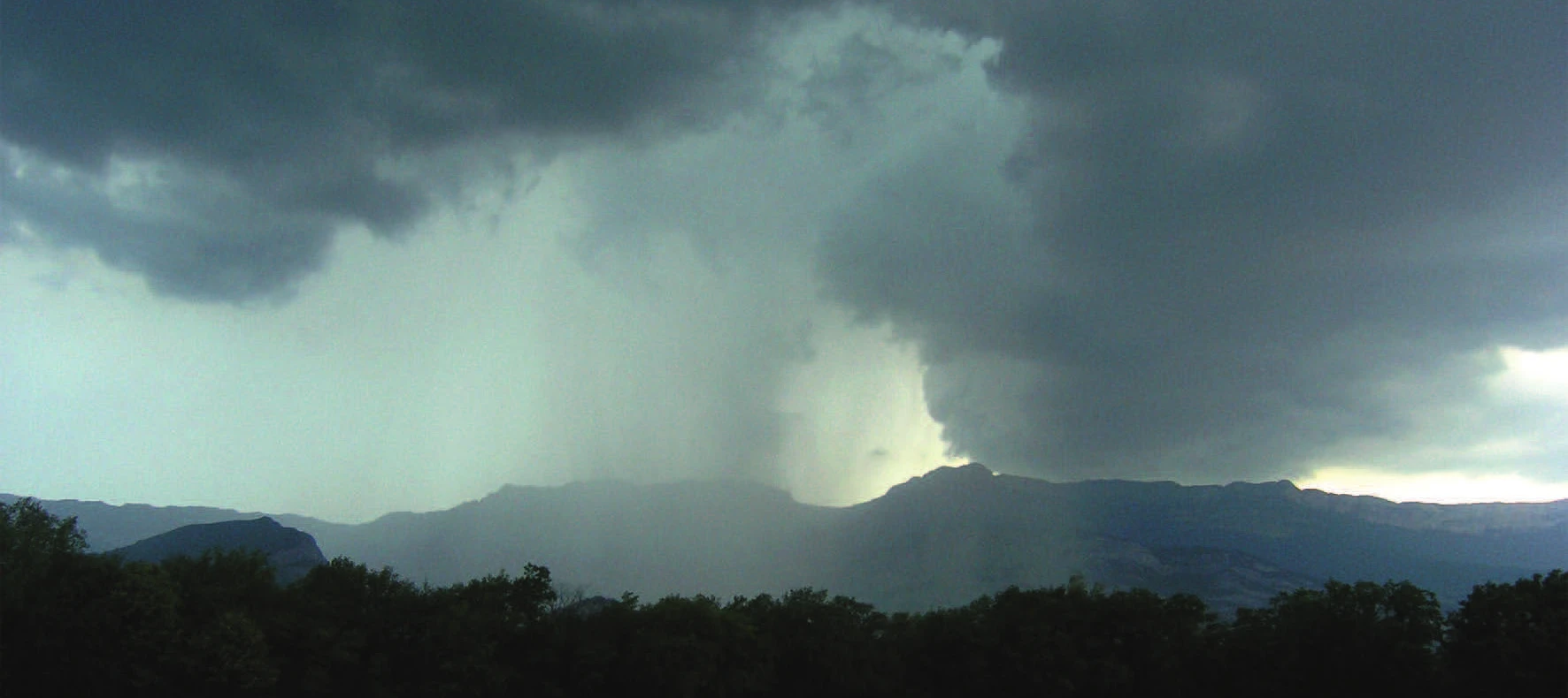 Cumulonimbus clouds building over a forested ridge, with rain visible beneath the cloud base