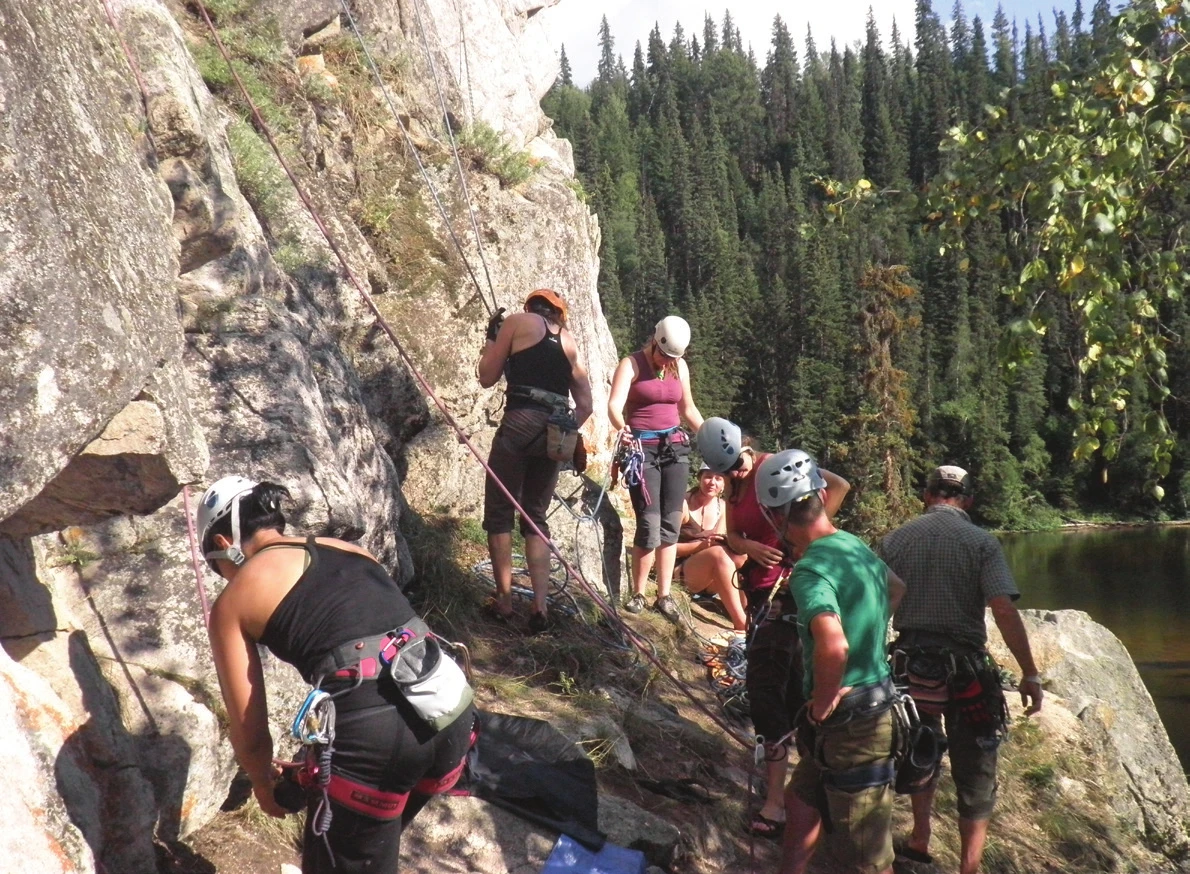Un groupe d’escalade en moulinette sur une falaise au bord d’un lac, les participants rassemblés au pied pendant que deux grimpeurs progressent sur des voies voisines