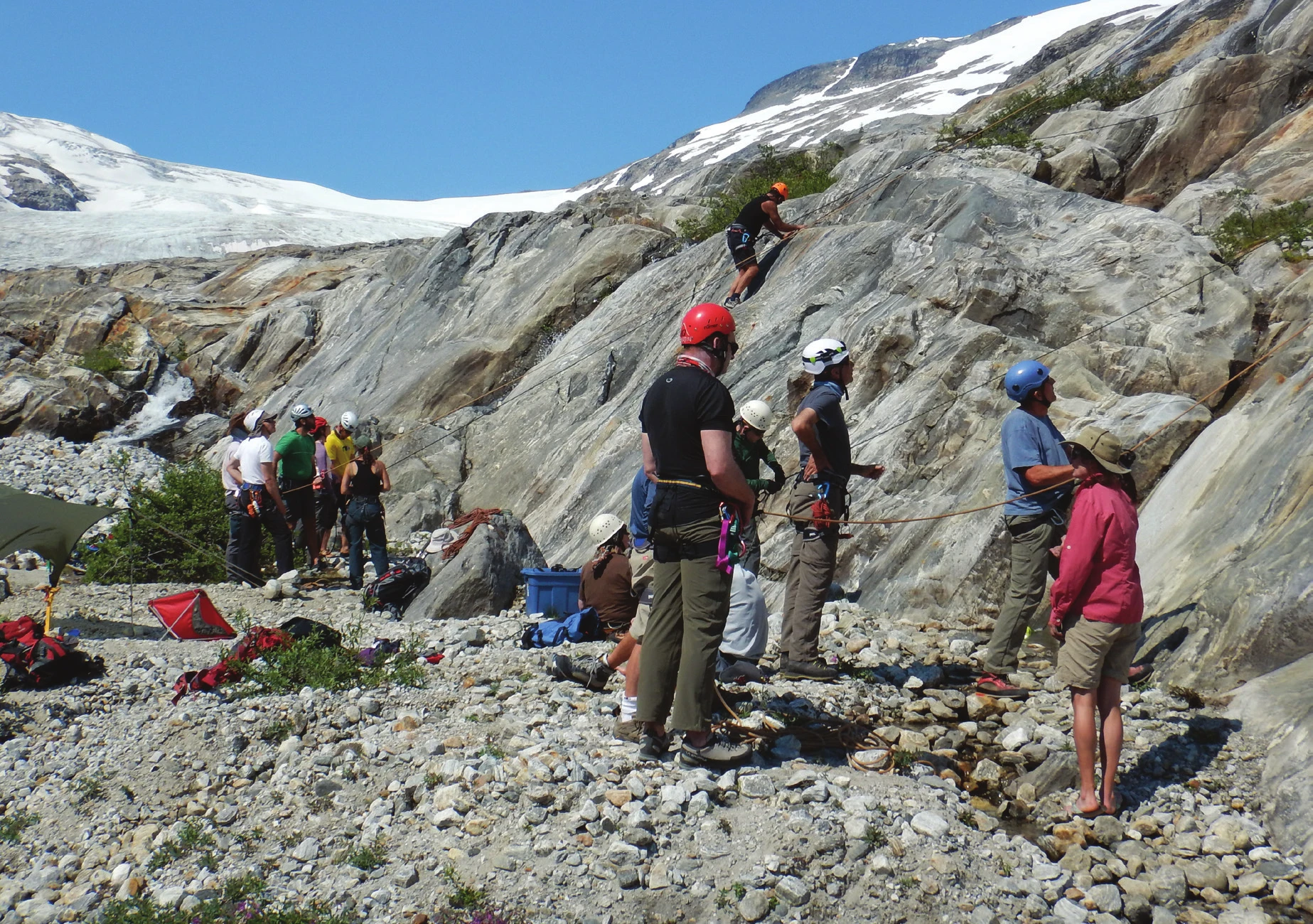 Un groupe d’escalade en moulinette rassemblé au pied d’une dalle alpine, un grimpeur haut sur la paroi et un glacier en arrière-plan